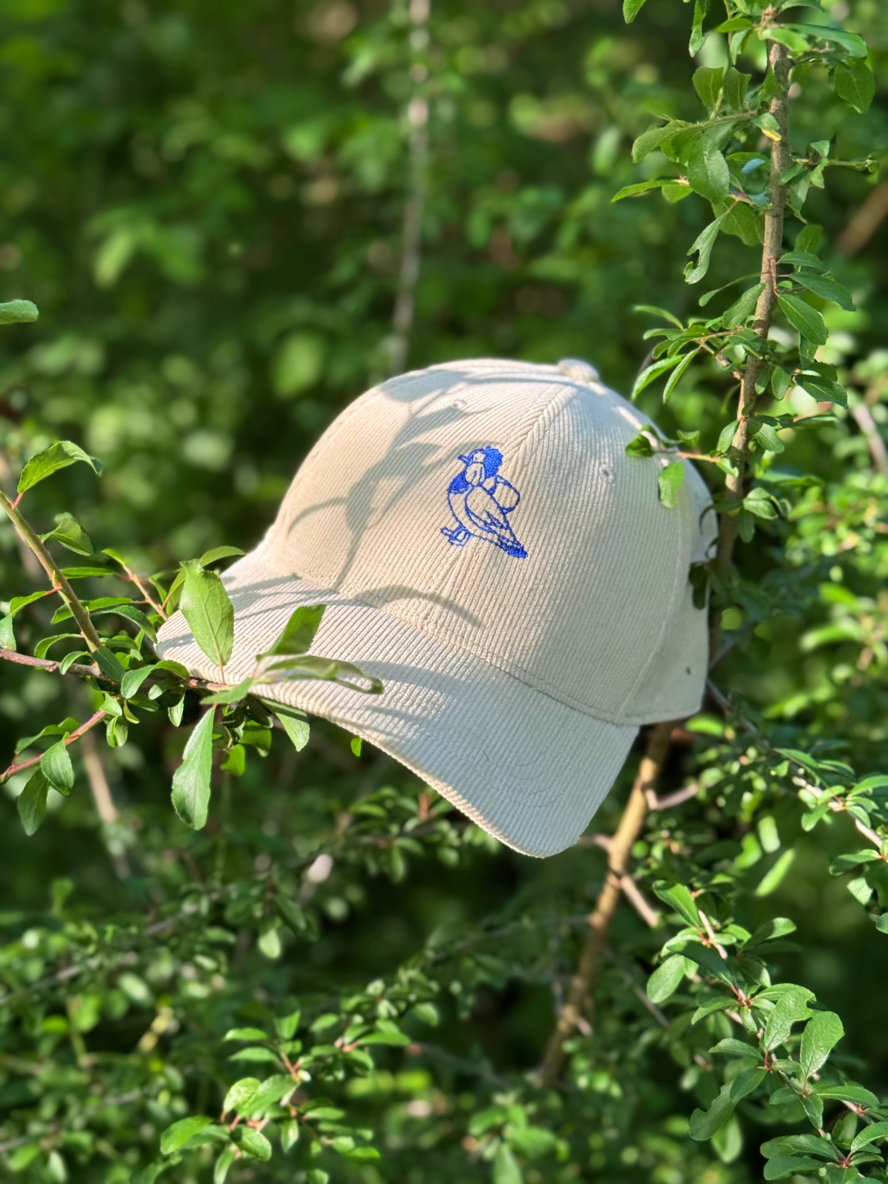 Photo of a natural white cord cap hanging lying on some branches of a plant. In the center of the cap is a stylised blue logo of a mandarin duck.